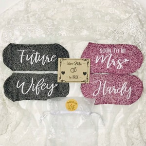 May include: Two pairs of socks, one gray and one pink, with white text. The gray socks say "Future Wife" and the pink socks say "Soon to be Mrs. Hardy". A small card with the text "Future Mrs. - the Bride" is in the center of the image.