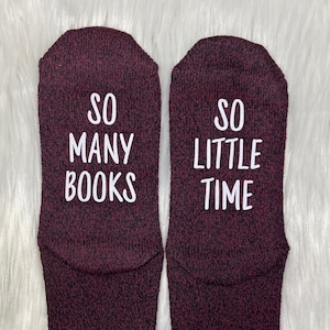 May include: A pair of burgundy socks with the text "SO MANY BOOKS" and "SO LITTLE TIME" in white block letters. The socks are made of a soft, textured material and are displayed on a white, fluffy surface. The socks are a cozy accessory for book lovers.