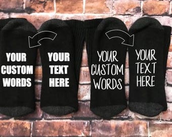 May include: Four black socks with gray heels and toes are displayed. Each sock has the words "YOUR CUSTOM WORDS" and "YOUR TEXT HERE" printed in white, with an arrow pointing to the text. The socks are on a brick background.
