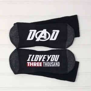 Puede incluir: Un par de calcetines negros con un estampado gráfico blanco. Un calcetín dice "DAD" con el logotipo de los Vengadores. El otro calcetín dice "I LOVE YOU THREE THOUSAND".