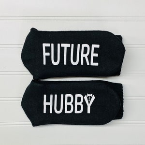 May include: A pair of black socks with white text. The top sock says "FUTURE" and the bottom sock says "HUBBY" with a small tuxedo graphic.