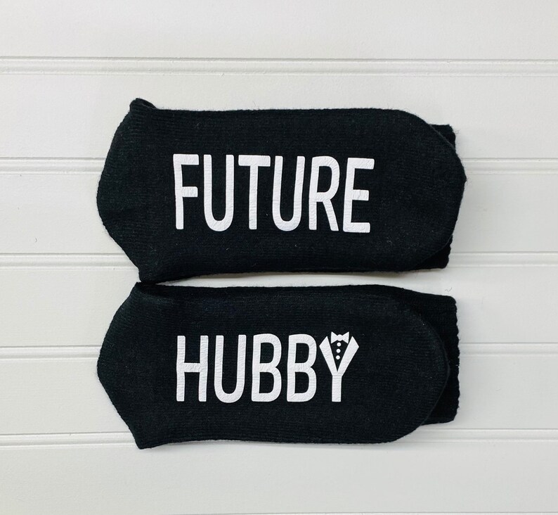 May include: A pair of black socks with white text. The top sock says "FUTURE" and the bottom sock says "HUBBY" with a small tuxedo graphic.