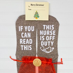 May include: A pair of gray socks with white text. One sock says "If you can read this" and the other sock says "This nurse is off duty". The socks are packaged in a red mesh bag with a brown tag that says "Handmade with love".