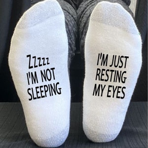 Peut inclure: Une paire de chaussettes blanches avec du texte noir. Une chaussette indique "Zzzzzz Je ne dors pas" et l'autre chaussette indique "Je ne fais que me reposer les yeux".