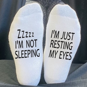 Puede incluir: Calcetines blancos con texto negro. Un calcetín dice "Zzzzz No estoy durmiendo" y el otro dice "Solo estoy descansando mis ojos".