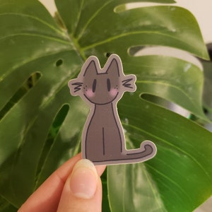 Puede incluir: Una pegatina de gato negro con mejillas rosadas y un contorno blanco. La pegatina está en una hoja verde.