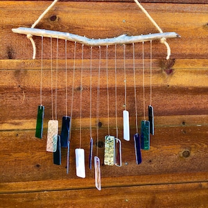 Peut inclure: Un carillon artisanal avec une branche de bois flotté suspendue par une corde naturelle. Diverses pièces de verre rectangulaires vertes, bleues, blanches et dorées pendent de la branche, créant un accent décoratif et mélodique.