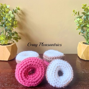 May include: Handmade crochet donuts in pink, white, and brown. The donuts are displayed on a dark wooden surface, flanked by two potted plants in geometric wooden planters. The text "Gray Pleasantries" is visible in the image.