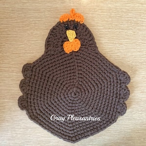 May include: Brown crocheted chicken-shaped hot pad with orange and yellow details for the beak and comb. The hot pad has a circular centre and a scalloped edge. The text "Gray Pleasantries" is visible in the centre of the hot pad.