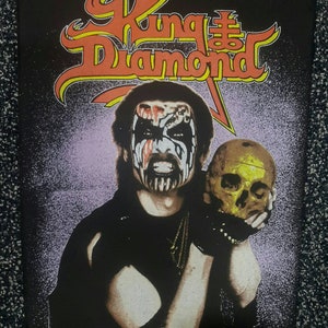 Pode incluir: Um pôster preto com a banda King Diamond. A imagem mostra uma pessoa com pintura facial segurando uma caveira. O nome da banda está em letras laranja e amarelas no topo. O fundo é um gradiente de roxo e branco.
