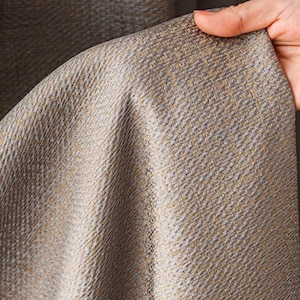 May include: A curtain panel featuring a textured fabric with a woven pattern in shades of grey and metallic gold. The material has a visible cross-hatch weave and a slight sheen, creating a dimensional appearance across the surface of the fabric.