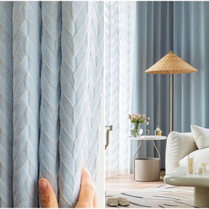 May include: Close-up of light blue curtains with a chevron pattern. The curtains are hung in a living room with a white couch, a side table, and a lamp with a woven shade.