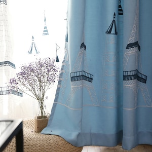 May include: Light blue curtains with a white and black embroidered design of the Eiffel Tower. The curtains are hung in a window with a white sheer curtain behind them.