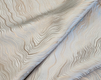 May include: Close-up of a folded fabric with a flowing, wave-like pattern. The fabric features a combination of light beige, silver, and gold tones, creating a textured, shimmering effect. The design resembles a topographical map.