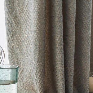 May include: Close-up of textured, light gray curtains with a subtle chevron pattern. The fabric drapes in soft folds, creating a sense of depth and texture. The curtains appear to be made of a woven material, adding a touch of elegance to any room.