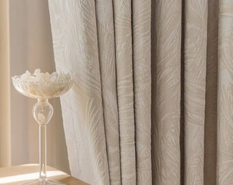 May include: Beige curtains with a subtle leaf pattern hang next to a wooden table. A clear glass pedestal bowl filled with white decorative stones sits on the table. The curtains are made of a textured fabric.