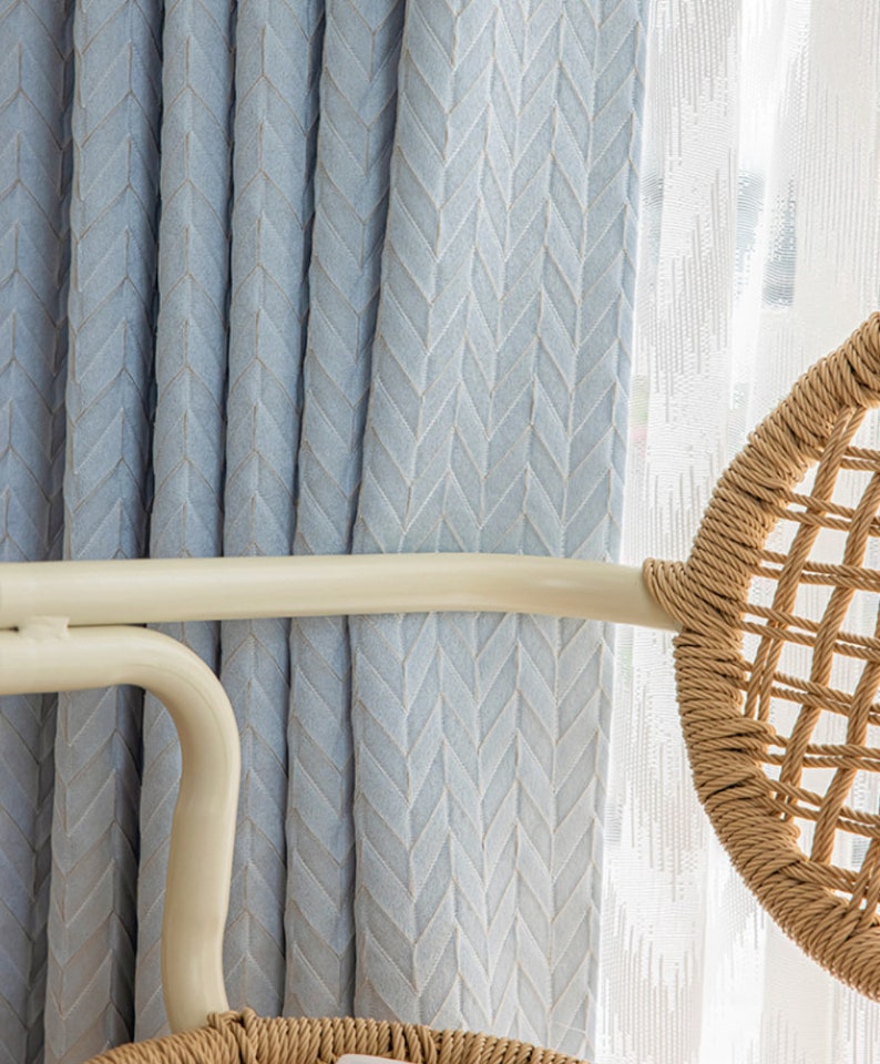 May include: Light blue curtains with a subtle herringbone pattern hang in front of a window. The curtains are partially drawn, revealing a white sheer curtain behind them. A wicker chair is partially visible in the background.