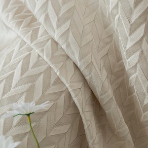 May include: Close-up of a beige fabric with a textured geometric pattern. The fabric is folded and has a soft, luxurious feel.
