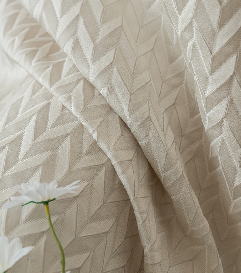 May include: Close-up of a beige fabric with a textured geometric pattern. The fabric is folded and has a soft, luxurious feel.