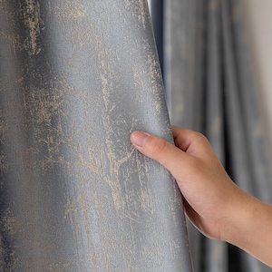 May include: Close-up of a textured, gray curtain with a distressed, mottled pattern in shades of beige and gray. The fabric has a subtle sheen, and the design evokes a sense of aged elegance. The curtain is held open, revealing a glimpse of another curtain.