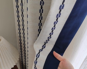 Könnte beinhalten: Cremefarbene Leinenvorhänge mit vertikalen Reihen dunkelblauer und schwarzer Stickereidetails. Ein marineblauer Rand verläuft entlang der Kante. Die Vorhänge sind teilweise geöffnet und zeigen die Textur und das Gewebe des Stoffes.