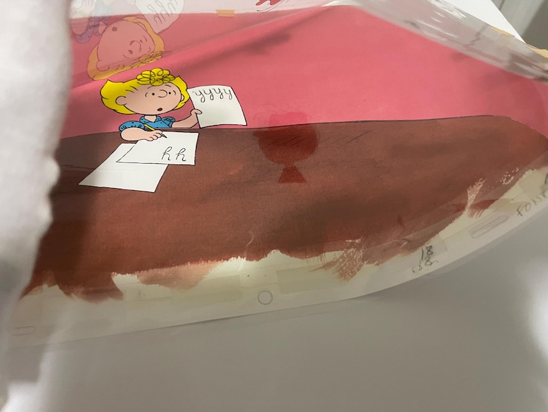 May include: A cartoon illustration of a girl with blonde hair sitting at a desk writing on a piece of paper. The paper has the letters "R.H." written on it. The girl is wearing a blue shirt and a red bow. The desk is brown and the background is pink.