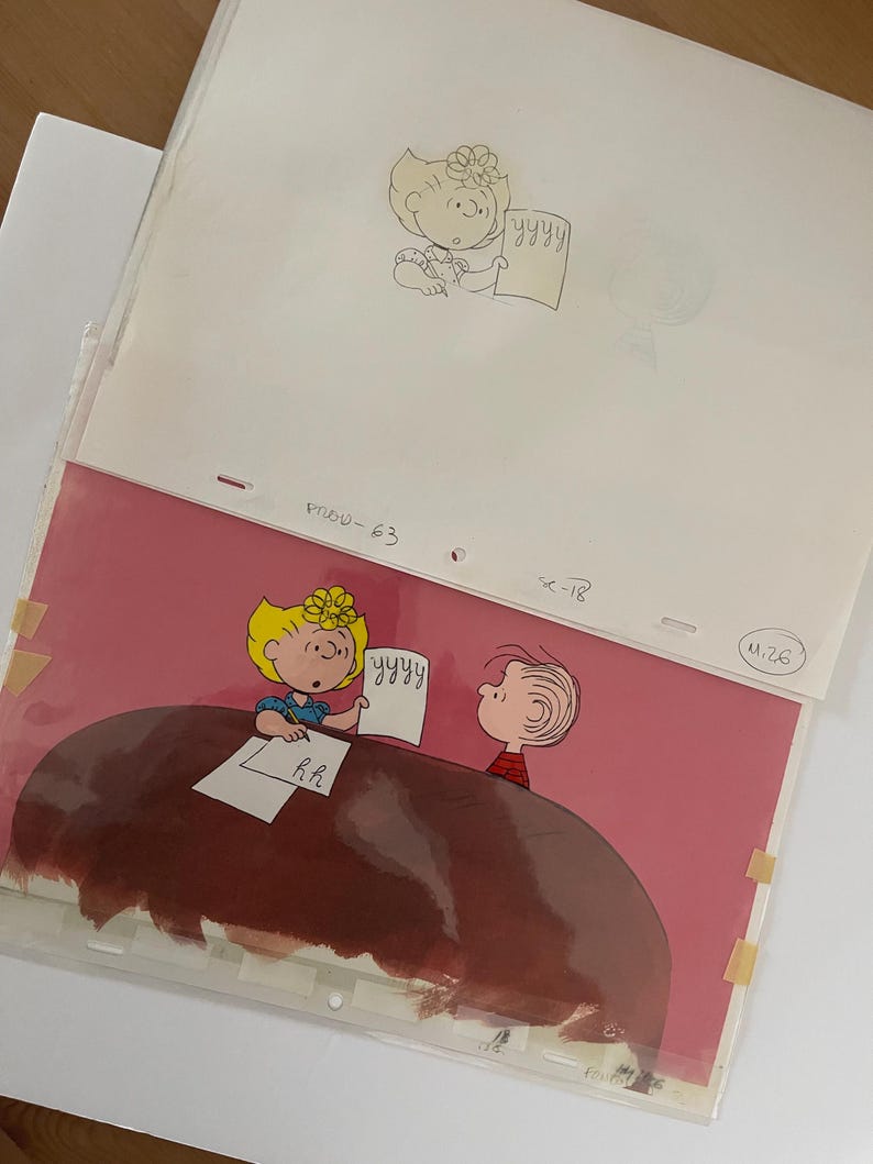 May include: A pink background with a brown table. Two cartoon characters are sitting at the table. One character is a girl with blonde hair and a blue shirt. The other character is a boy with brown hair and a red shirt. The girl is holding a piece of paper with the word "YYYYY" written on it. The boy is looking at the girl. The text "R.H." is written on the table.