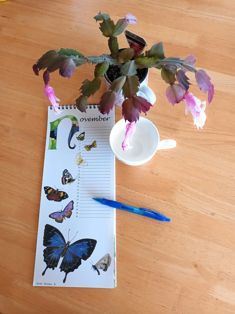 K&ouml;nnte beinhalten: Eine Kalenderseite f&uuml;r November mit einer blauen Schmetterlings-Illustration und einem blauen Stift auf einem Holztisch. Der Kalender hat einen wei&szlig;en Hintergrund mit schwarzen Zahlen und einem schwarzen Rand. Der Schmetterling befindet sich in der unteren rechten Ecke der Seite. Der Stift liegt auf der Seite und der Tisch ist aus Holz.