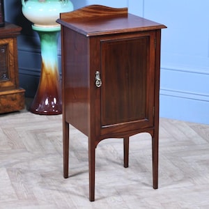 May include: A small, antique wooden cabinet. It has a single door and a curved top. The cabinet is made of dark wood and has a silver-coloured handle. It stands on four slender legs. The cabinet is likely used for storage.