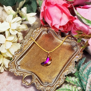 May include: A gold chain necklace with a crescent moon pendant made of iridescent purple glass. The necklace is displayed on a decorative brass tray with a floral background.