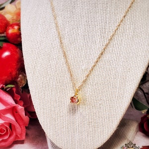 May include: A delicate gold chain necklace with a small, square-shaped pendant. The pendant is filled with red glitter and has a gold border.
