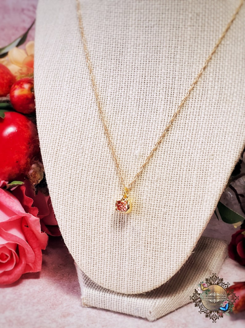 May include: A delicate gold chain necklace with a small, square-shaped pendant. The pendant is filled with red glitter and has a gold border.