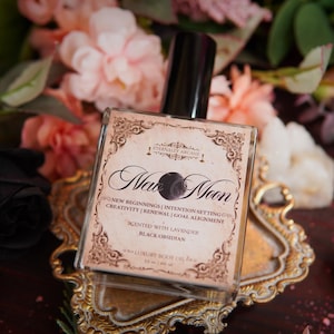 May include: A square glass bottle of luxury body oil with a black cap, labeled "New Moon." The label features ornate scrollwork and the words "New Beginnings, Intention Setting, Creativity, Renewal, Goal Alignment." The bottle is on a gold-colored decorative tray, surrounded by pink and black flowers.