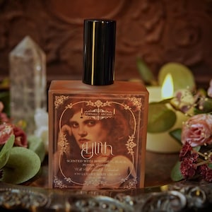 May include: A square perfume bottle with a black cap and a label featuring a woman's portrait. The label reads "Ylth" and "Scented with Bergamot, Black Pepper, Jasmine". The bottle is surrounded by flowers and a lit candle.