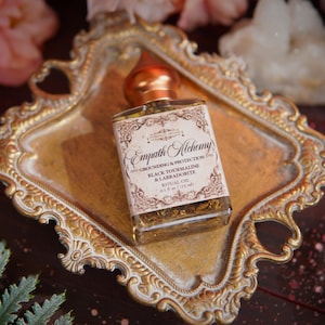 May include: A square glass bottle of ritual oil with a copper-colored cap sits on a gold-colored ornate tray. The label reads "Empath Alchemy" and lists ingredients. The tray is surrounded by pink flowers and greenery, creating a mystical aesthetic.