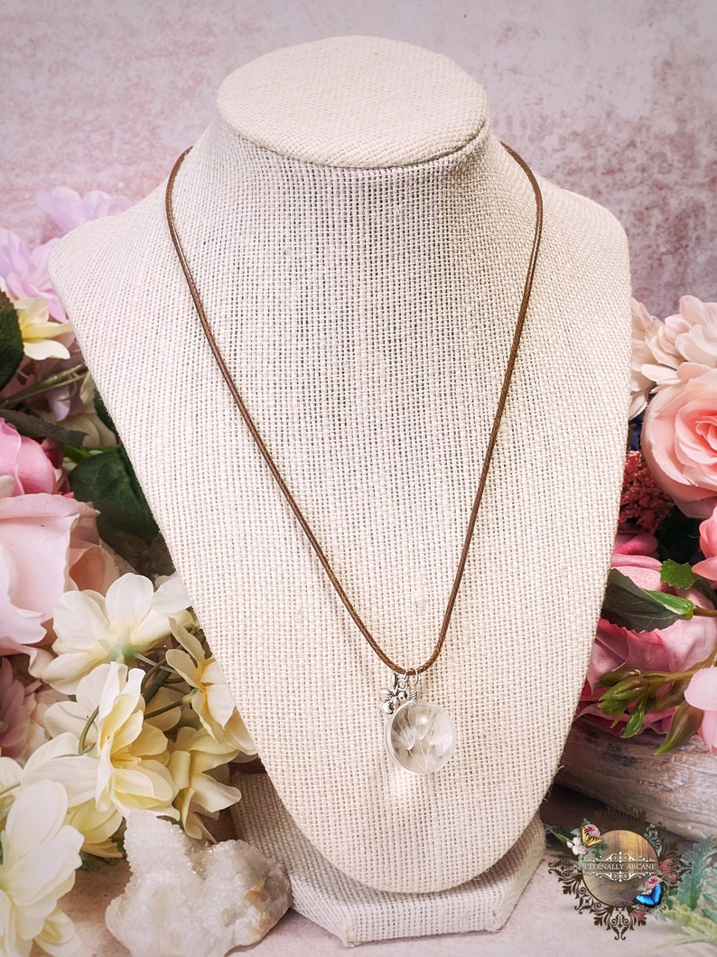 May include: A brown leather cord necklace with a silver pendant featuring a clear glass orb containing a dried dandelion. The pendant has a small silver flower charm attached.