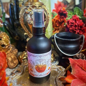 Może przedstawiać: Czarna szklana butelka z czarną dyszą rozpylającą i białą etykietą z napisem "Witchcraft in Autumn" ze stylizowanym wzorem pomarańczowej dyni. Butelka stoi na złotej powierzchni z jesiennymi liśćmi i czarnym kotłem.