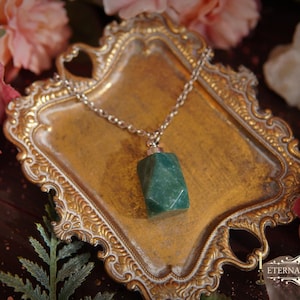 May include: A silver chain necklace with a rectangular, faceted, green gemstone pendant rests on a gold-colored, ornate tray. The tray is surrounded by pink roses and other floral elements. The text "Eternally Arcane" is visible.