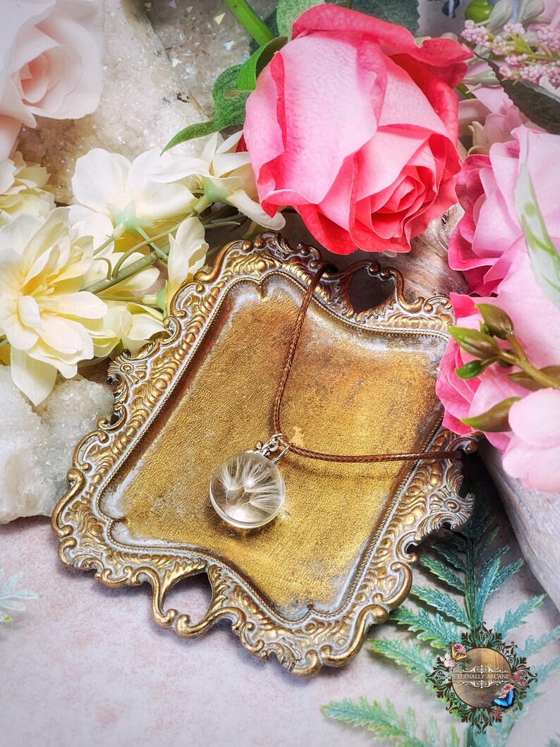 May include: A delicate necklace with a glass pendant containing a dried dandelion. The necklace is displayed on a gold-colored tray with a floral background.