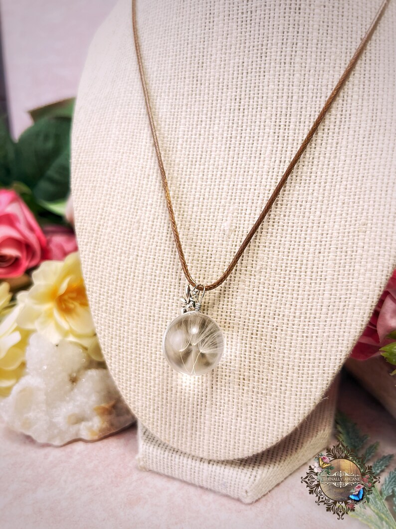 May include: A delicate necklace with a brown cord and a clear glass pendant containing a dried dandelion. The pendant is attached to the cord with a silver clasp.