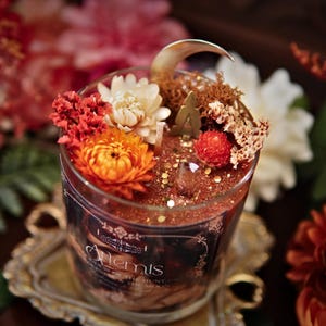 May include: A glass candle jar with a gold glitter top and a black label that reads "Alchemists" and "The Hunt". The jar is filled with a red and gold wax and decorated with dried flowers, including white, orange, and red flowers.