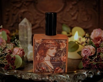 Puede incluir: Un frasco de perfume cuadrado, color ámbar, con tapón negro. La etiqueta presenta un retrato de mujer y el nombre "Persephone". El frasco está rodeado de flores, velas y un cristal sobre una bandeja plateada.