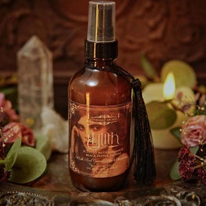 May include: A brown glass spray bottle with a black tassel, labeled "Lilith" with a woman's portrait. The bottle is surrounded by flowers, candles, and crystals. The label says "Scented with Bergamot, Black Pepper, Jasmine, With Red Garnet & Vanilla."