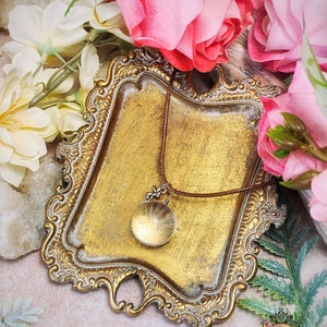 May include: A gold-toned ornate tray with a brown leather cord necklace with a clear glass pendant containing a dried dandelion. The tray is surrounded by white and pink flowers.