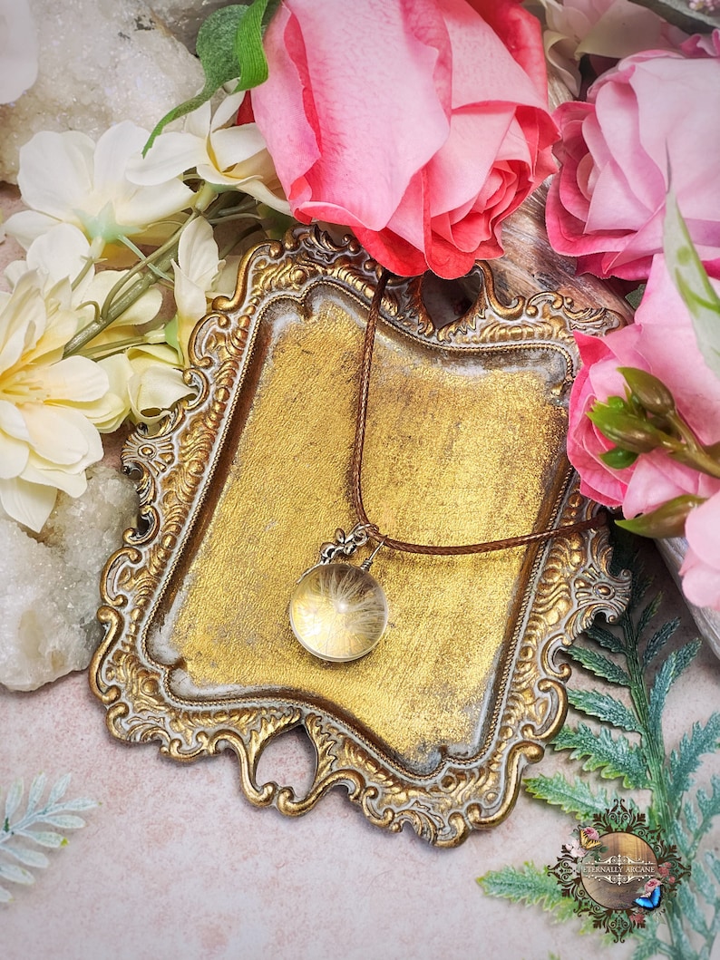 May include: A gold-toned ornate tray with a brown leather cord necklace with a clear glass pendant containing a dried dandelion. The tray is surrounded by white and pink flowers.