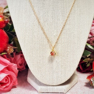 May include: A gold chain necklace with a small red and gold pendant shaped like a pomegranate. The necklace is displayed on a beige fabric mannequin.