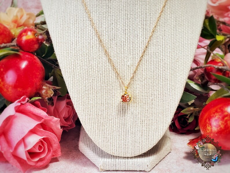 May include: A gold chain necklace with a small red and gold pendant shaped like a pomegranate. The necklace is displayed on a beige fabric mannequin.