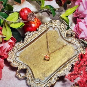 May include: A gold-tone necklace with a small, red heart-shaped pendant. The necklace is displayed on a decorative, ornate, gold-colored tray. The tray is surrounded by red and pink flowers.