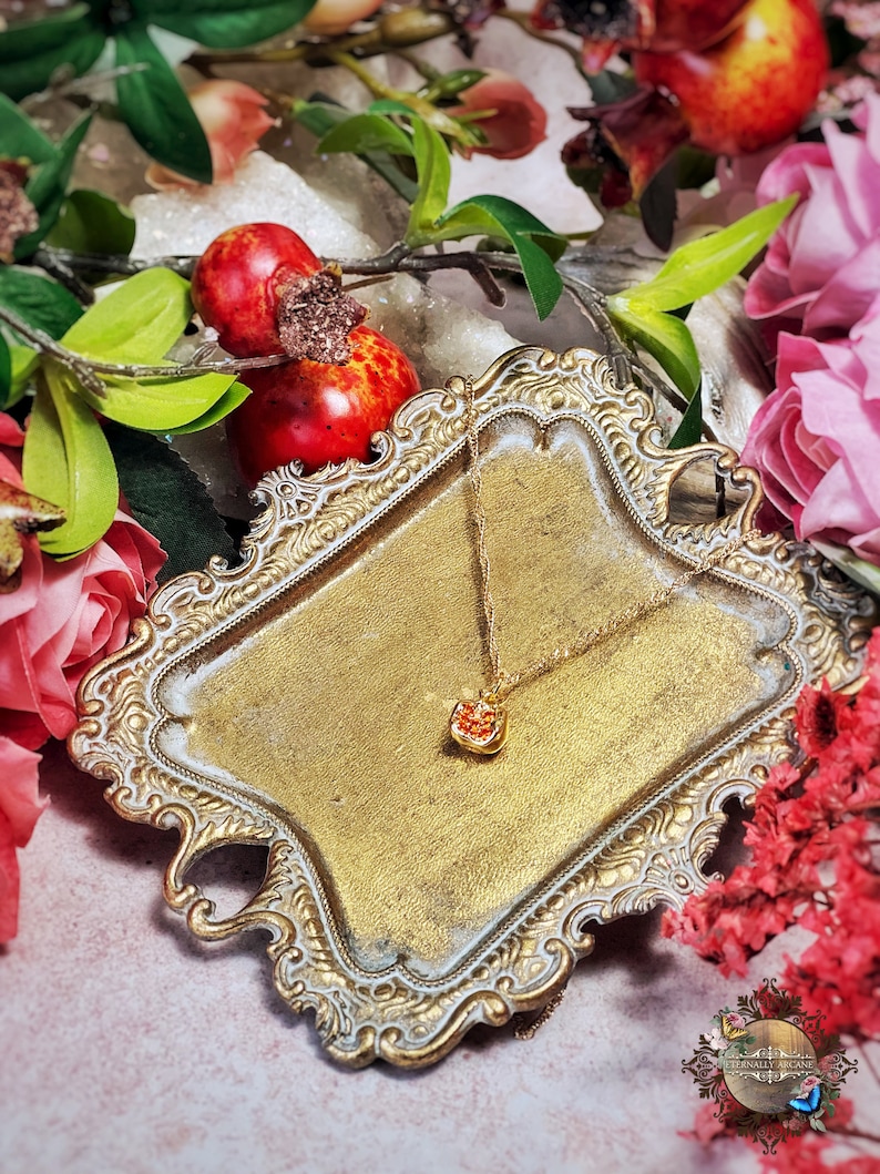 May include: A gold-tone necklace with a small, red heart-shaped pendant. The necklace is displayed on a decorative, ornate, gold-colored tray. The tray is surrounded by red and pink flowers.