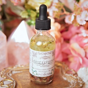 May include: A clear glass bottle with a black dropper top filled with a yellow liquid and rose petals. The label reads "Heart Coherence" and "Ritual Body Oil".